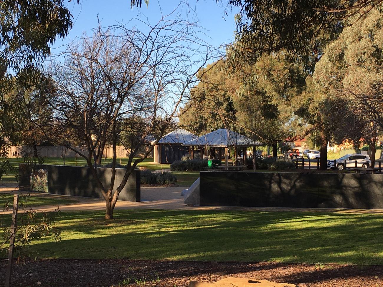 Green grass and trees with picnic shelters in the background and the bushfire memorial wall in the foreground