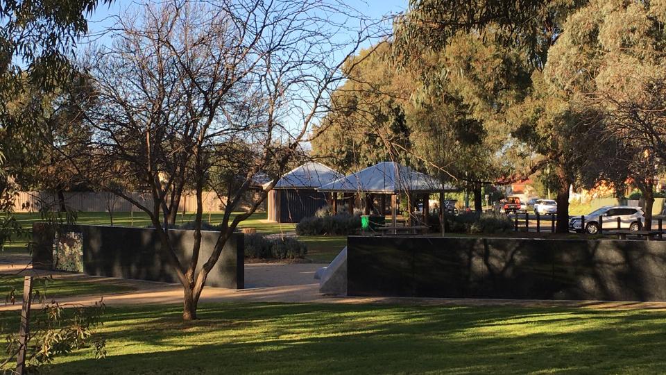 Green grass and trees with picnic shelters in the background and the bushfire memorial wall in the foreground