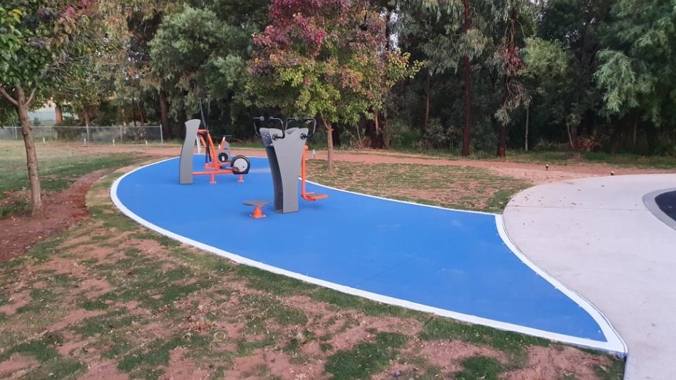 3 pieces of outdoor fitness equipment on blue paving with trees in the background