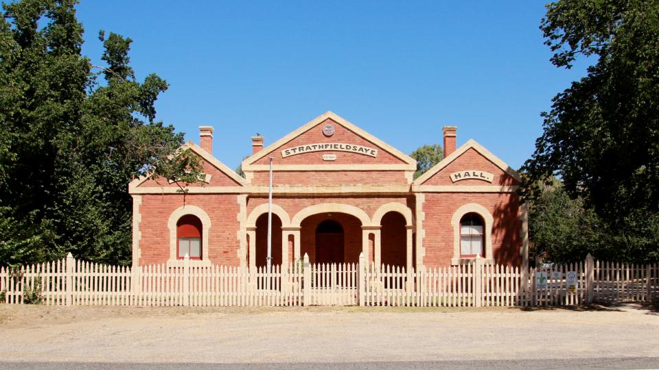 The Historic Strathfieldsaye Shire Building has a brick facade and many shady trees.